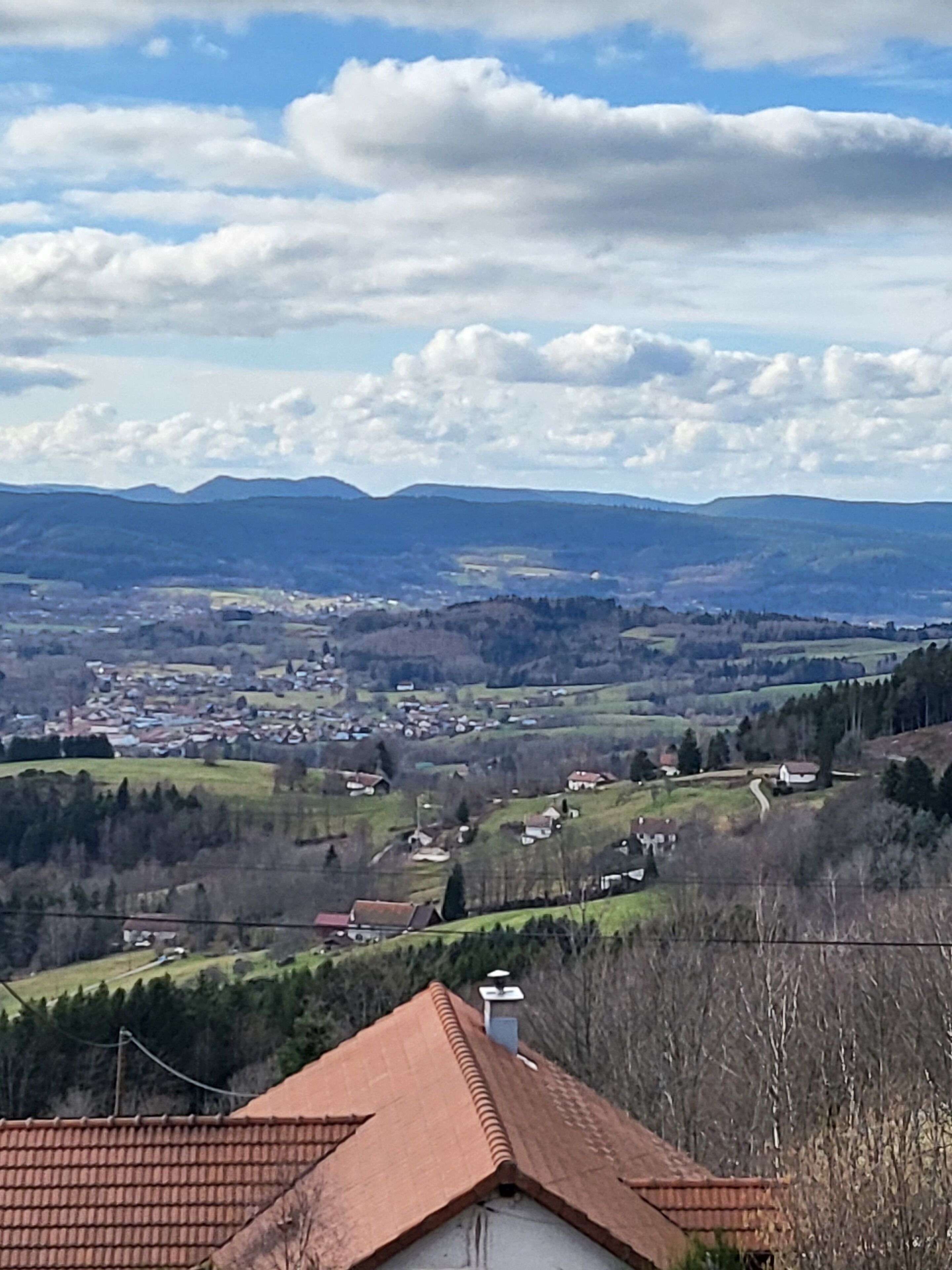 A rural landscape with a house featuring a red roof and a chimney, surrounded by hills and a village in the distance.