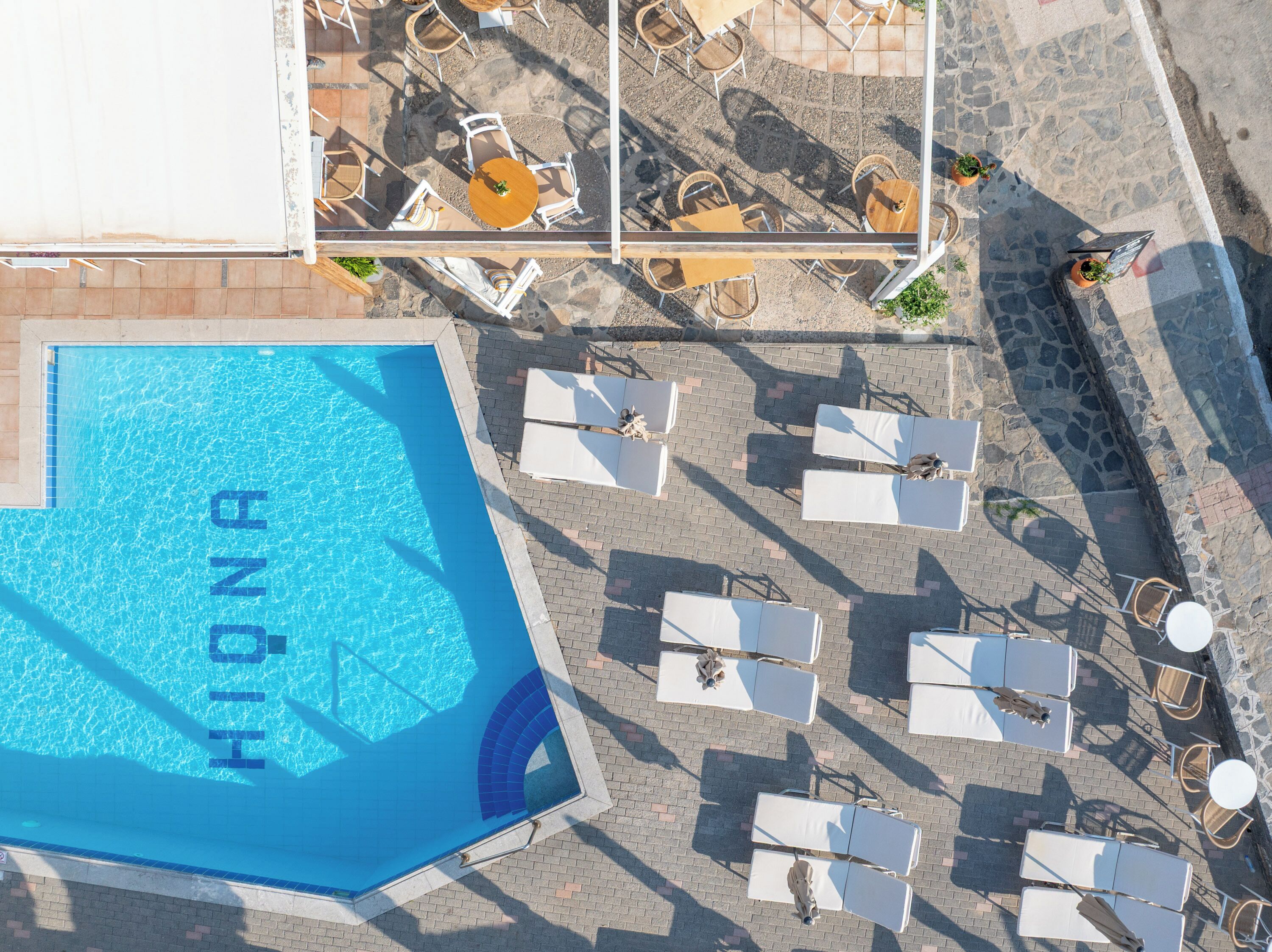 Vista aérea de un área de piscina de hotel con una piscina azul cristalina, tumbonas y mobiliario de exterior.