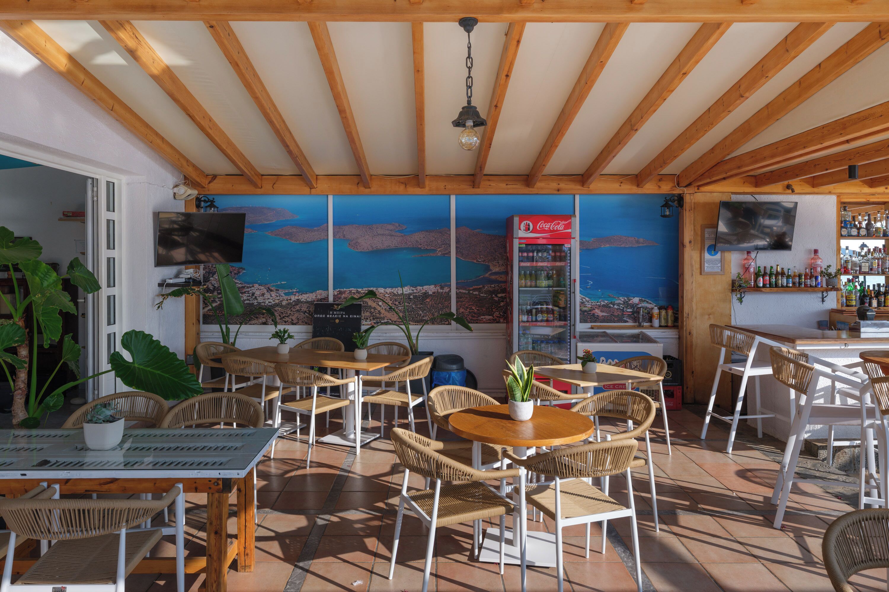 En restaurant med træbjælker, fletstole, et Coca-Cola køleskab og et muralmaleri af et kystlandskab.