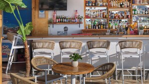 Een bar met houten balken, rieten stoelen en diverse flessen op planken.