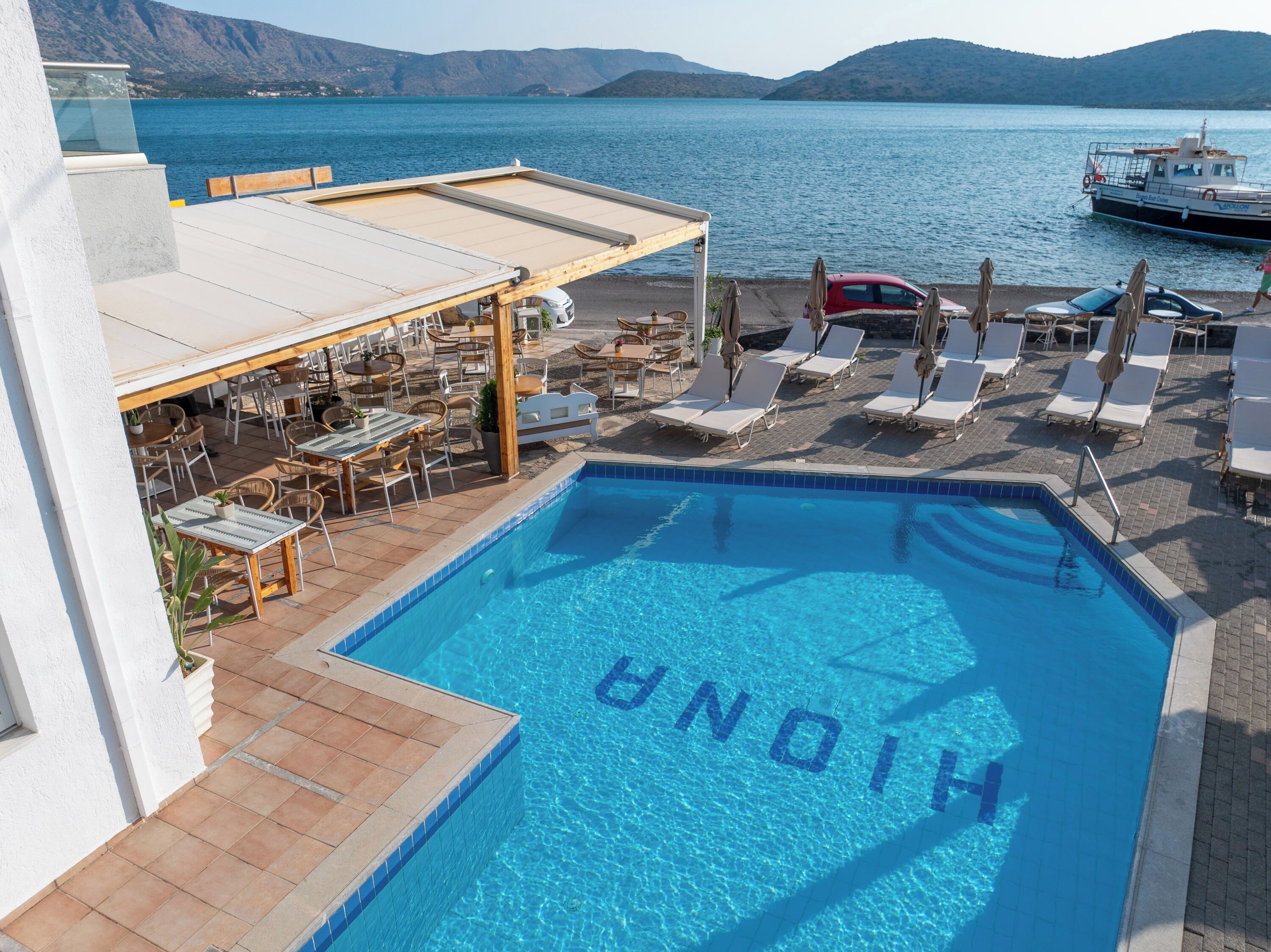 Una piscina con la palabra "HINON" escrita en el agua, rodeada de sillones de descanso y mesas, con vista al mar y a barcos al fondo.
