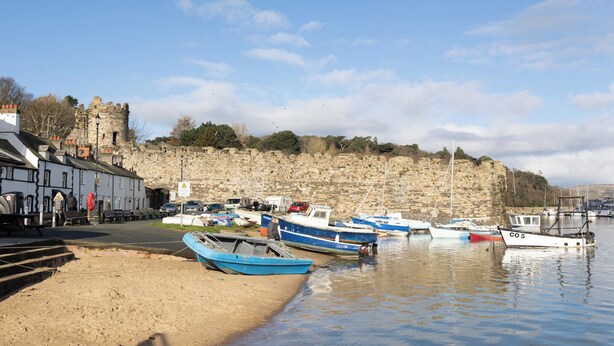 A coastal town with a historic stone wall, boats docked along the shore, and a building with a white facade. - Water View Cottage, Water View Cottage (Conwy)
