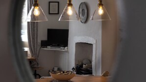 A view through a circular window into a room with a wooden table, hanging pendant lights, and a fireplace. - Water View Cottage, Water View Cottage (Conwy)