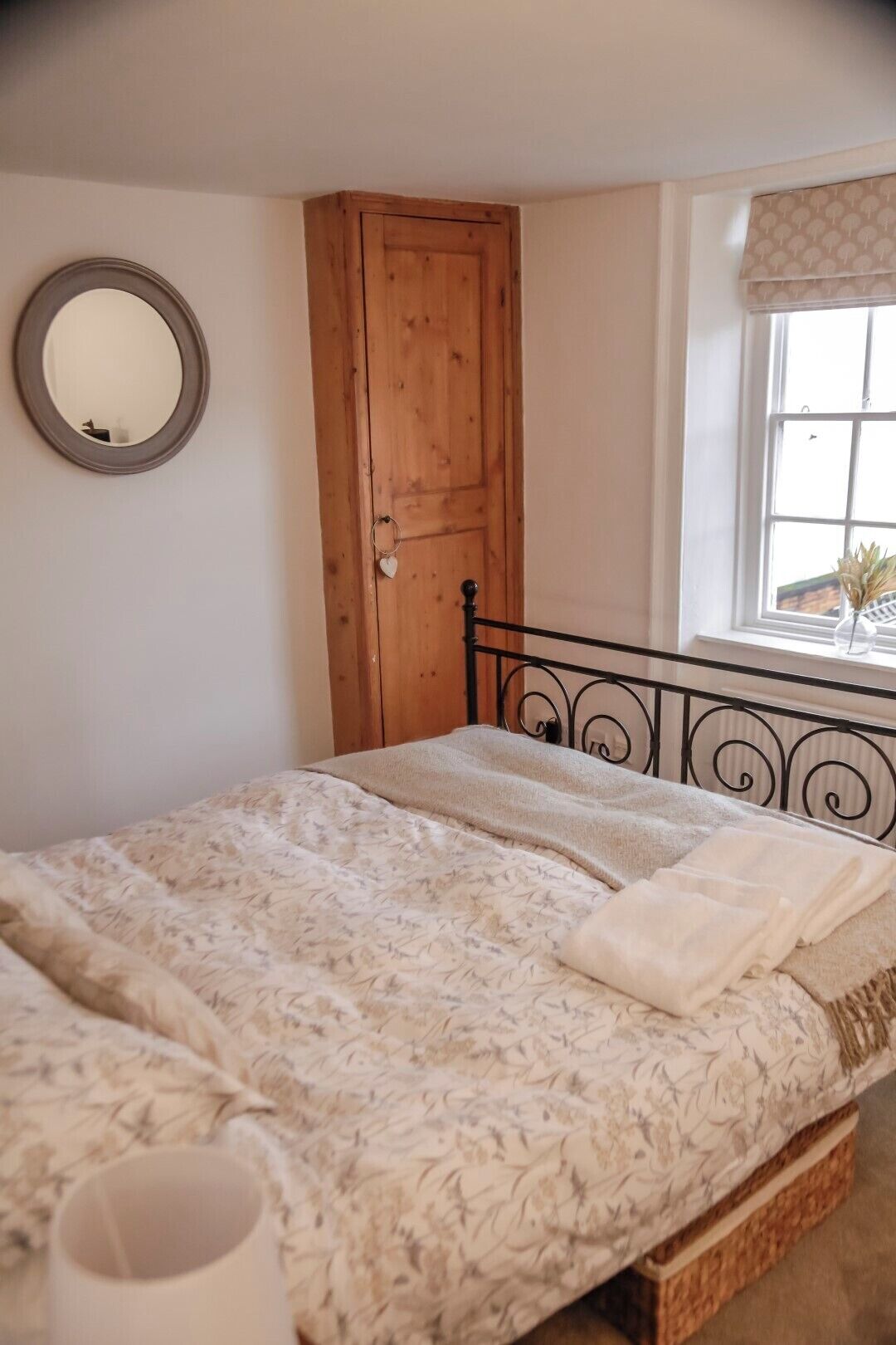 A bedroom with a bed, a wooden wardrobe, a round mirror, and a window with a view.