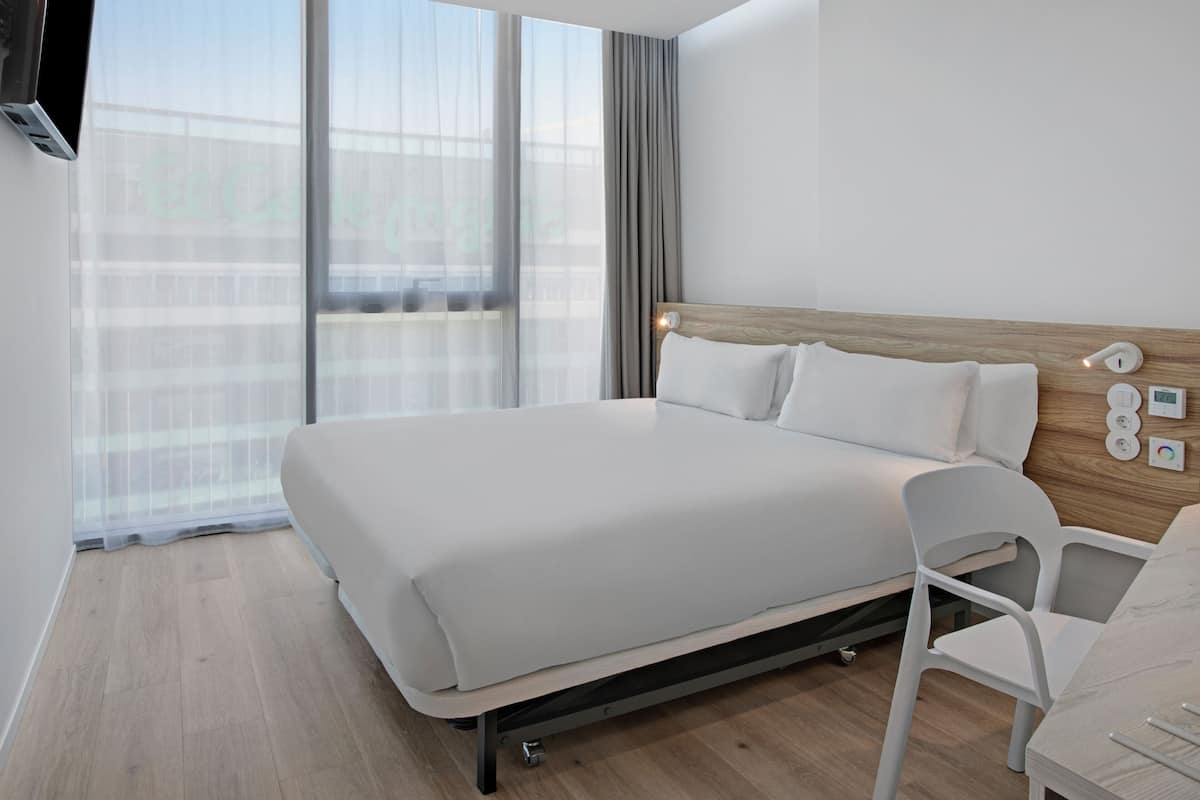 A modern hotel room with a large bed, a wooden headboard, a white chair, and a large window with sheer curtains.