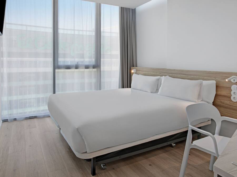 A modern hotel room with a large bed, a wooden headboard, a white chair, and a large window with sheer curtains.