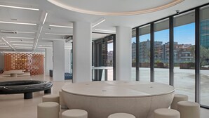 A modern interior with a circular seating area surrounded by cylindrical stools, large windows offering a view of buildings and greenery outside. - B&B Hotel Porto Gaia (Vila Nova de Gaia)