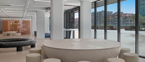 Ein modernes Interieur mit einer runden Sitzecke, umgeben von zylindrischen Hocker, große Fenster mit Blick auf Gebäude und Grünflächen.