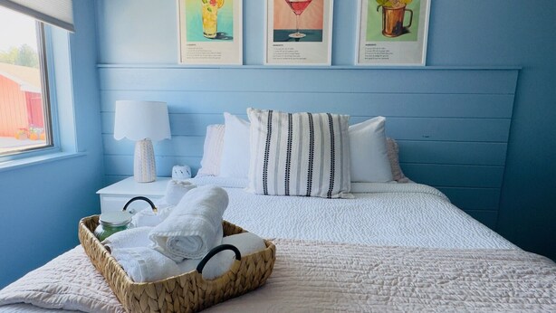 Un lit bien fait, avec une couette blanche et des oreillers, un panier rempli de serviettes pliées et une table de chevet avec une lampe. Trois photos encadrées de cocktails sont accrochées au mur.