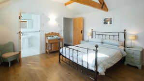 Een slaapkamer met een houten plafond, een metalen bedframe, een nachtkastje met lampen, een houten bankje, en een badkamer die door een open deur zichtbaar is.