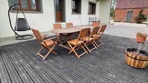En udendørs træterrasse med et rundt bord og stole, en hængekøjsstol og et lille haveområde med plantekrukker.