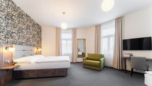 A hotel room with a bed, a green armchair, a desk with a chair, a TV, and a large window with curtains. - EA Hotel Victoria (Prague)