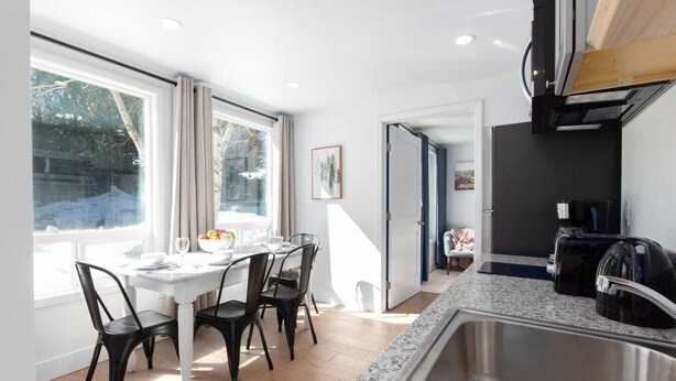 A modern kitchen with a stainless steel sink, a dining area with a white table and black chairs, and a view of the outdoors through large windows. - Downtown Mammoth | Walk to Shops & Eats! MH#5 (Mammoth Lakes)