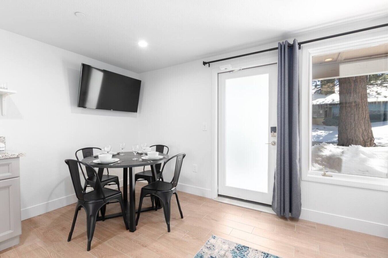 A dining area with a table set for four, a flat-screen TV mounted on the wall, and a sliding glass door leading to a snowy outdoor area.