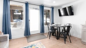 Ein Essbereich mit Tisch für vier Personen, einem an der Wand montierten Flachbildfernseher und einer Glasschiebetür mit blauen Vorhängen.