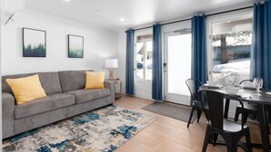 Ein modernes Wohnzimmer mit einem grauen Sofa, gelben Kissen, einem kleinen Tisch mit einer Lampe sowie einem Essbereich mit Stühlen und einem für zwei Personen gedeckten Tisch.