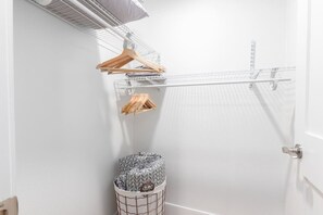 Une pièce blanche avec un présentoir métallique portant des cintres en bois, une poubelle à motifs et une porte blanche.
