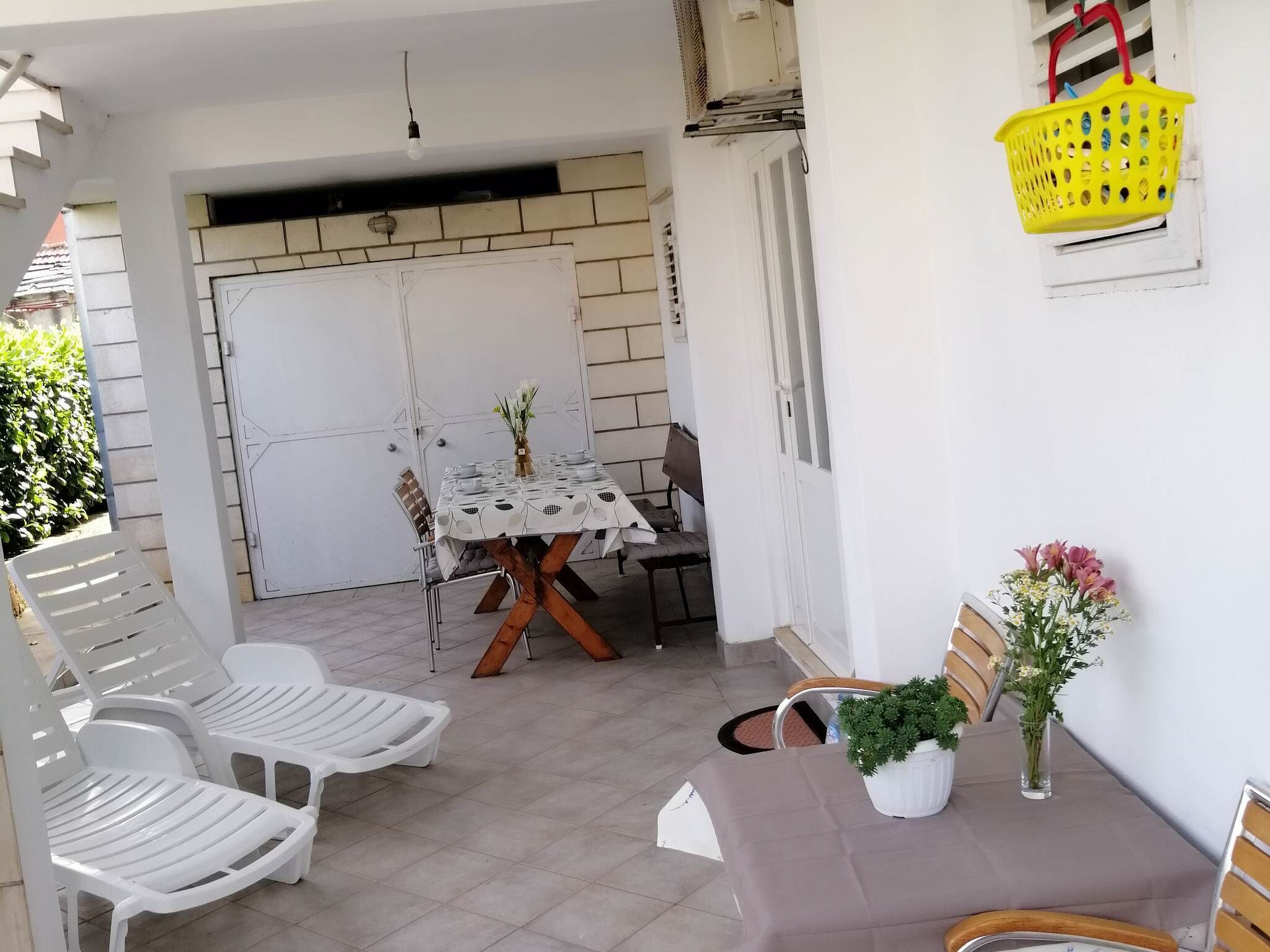 A patio area with a table set for two, surrounded by white plastic chairs. There is a yellow basket hanging on the wall and a potted plant on the table.