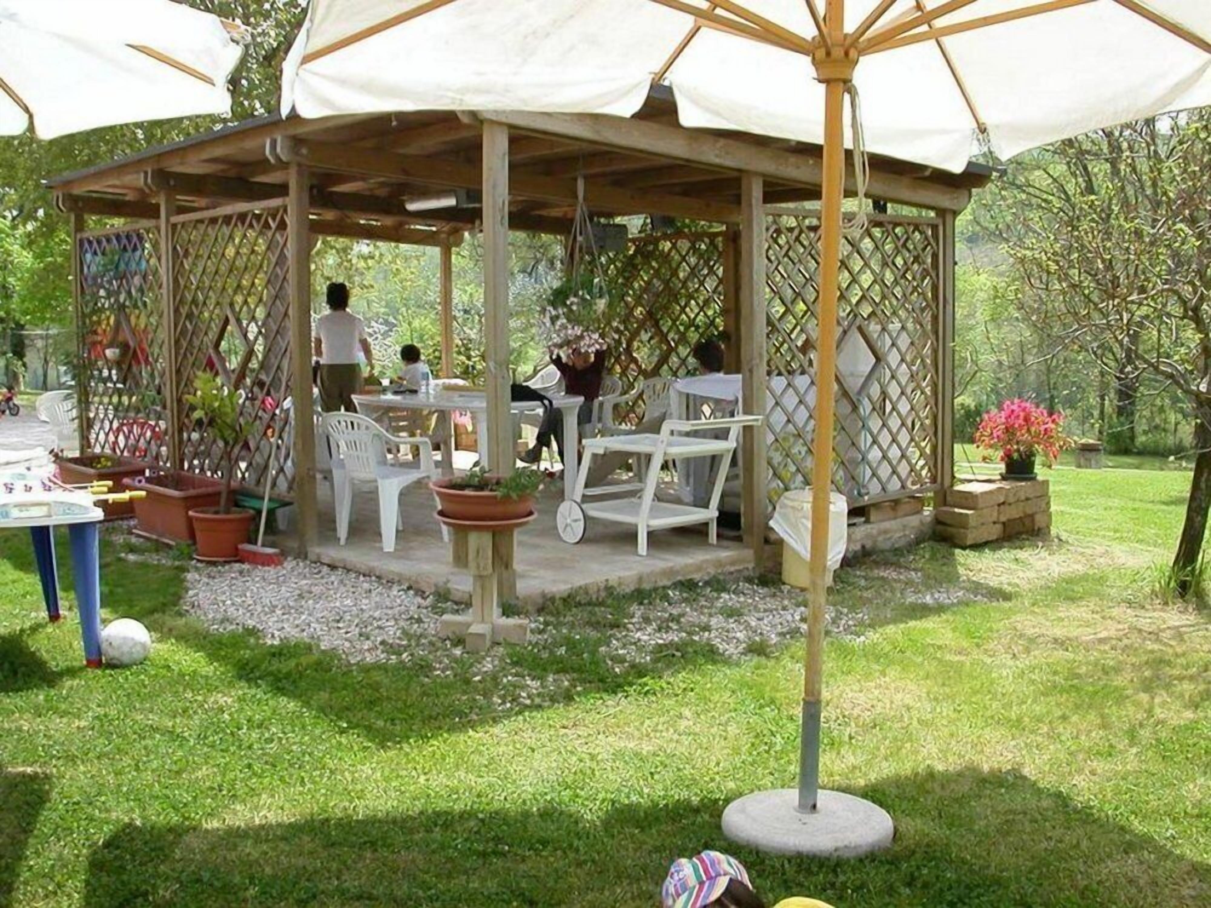 A covered outdoor seating area with tables and chairs, surrounded by greenery and a fence.