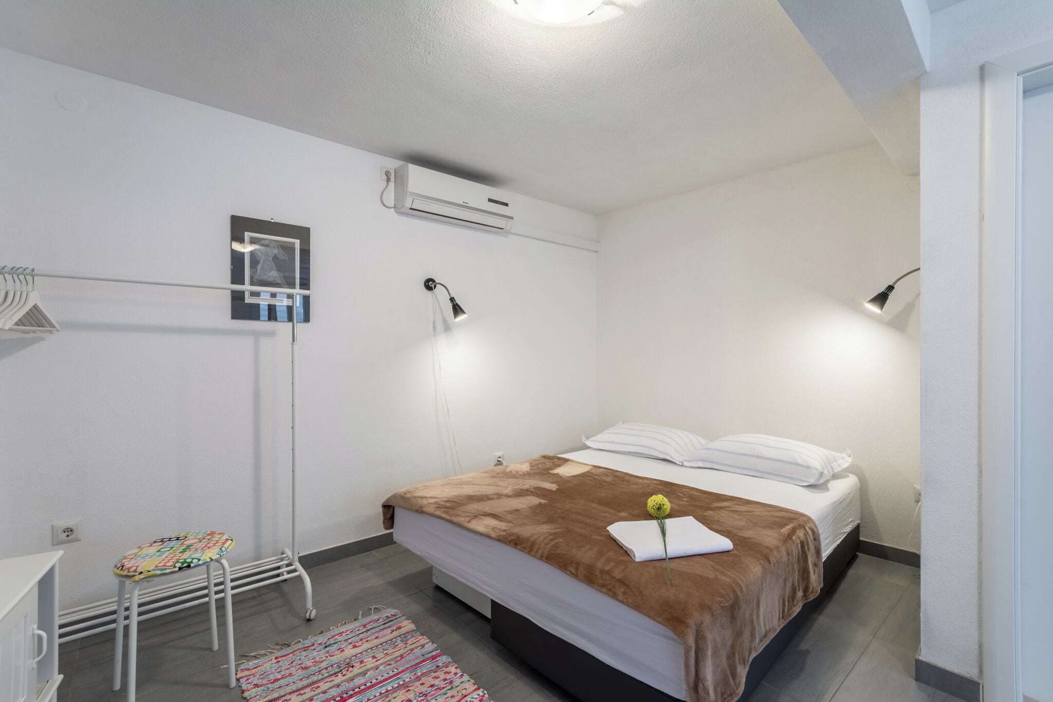 Een slaapkamer met een bed, nachtlampjes, een klein tafeltje, een stoel, een vloerkleed en een airconditioning.