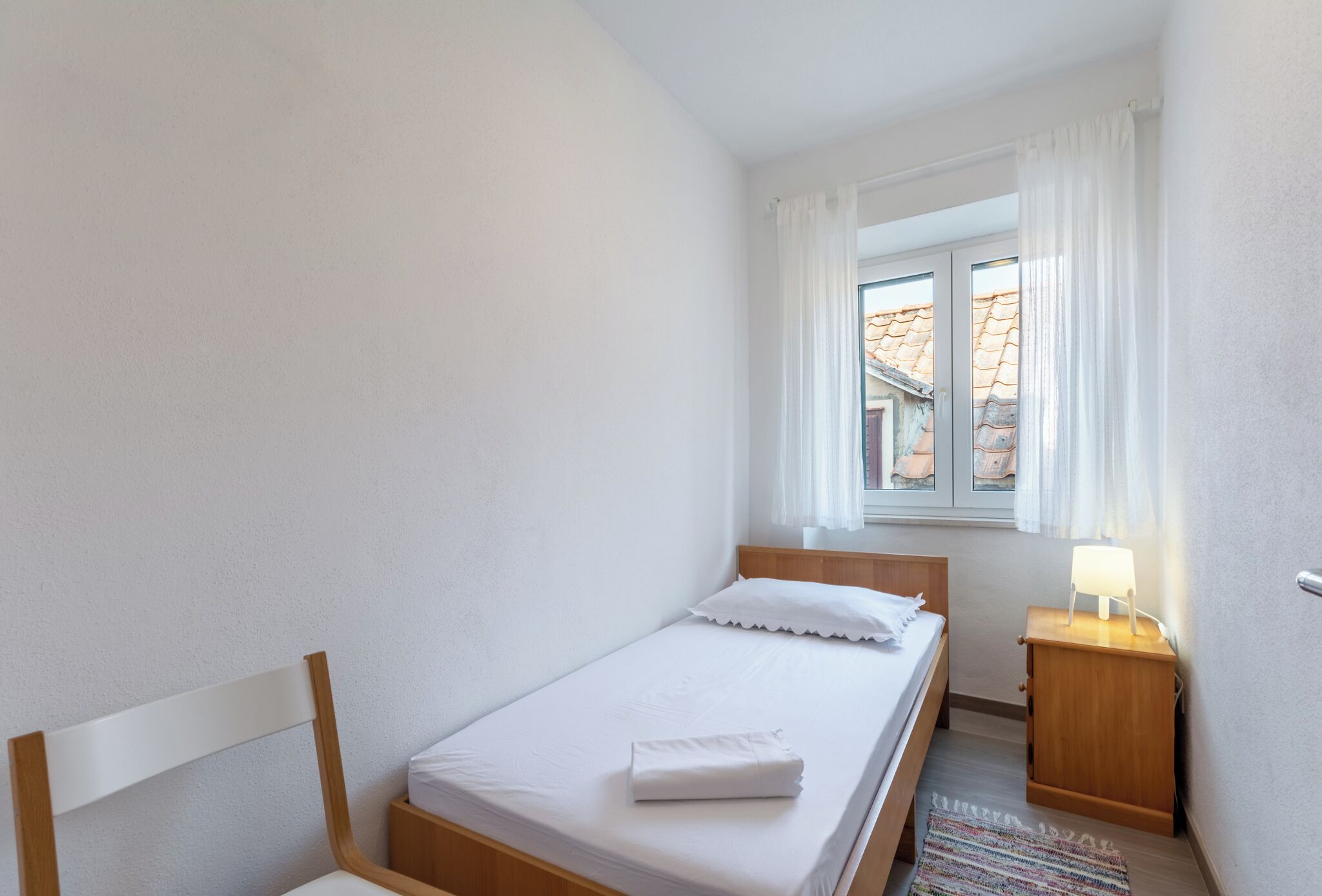 A small, neatly arranged bedroom with a single bed, a nightstand with a lamp, and a window with curtains.