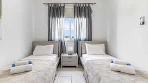 A hotel room with two single beds, a nightstand with a lamp, a window with curtains, and a view of the outdoors. - Iris Village 201 (Paphos)