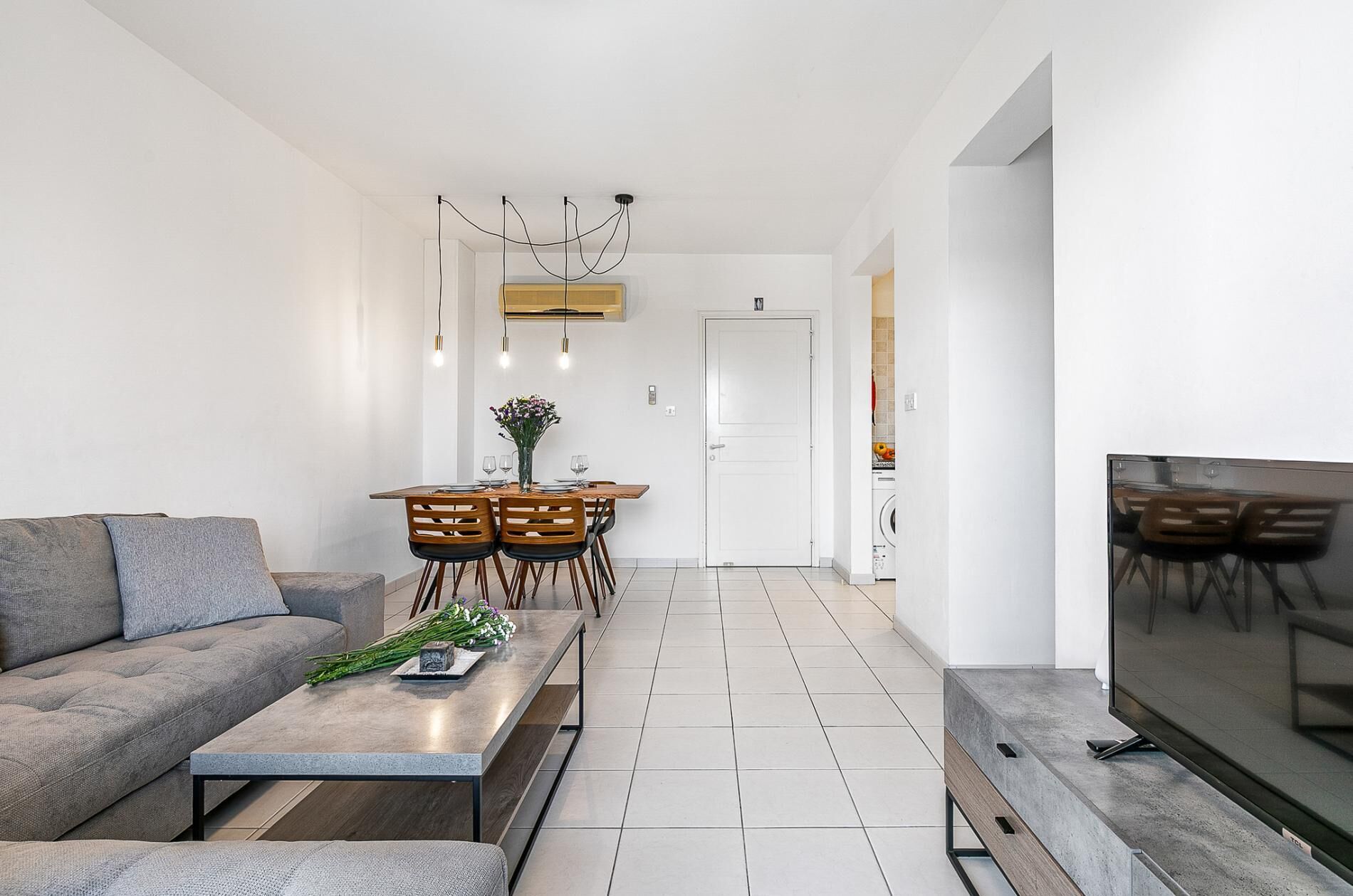 Una sala de estar moderna con sofá, mesa de centro y televisor. Hay un comedor con una mesa y sillas. La habitación tiene paredes blancas y piso de baldosas.
