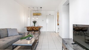 A modern living room with a sofa, coffee table, and TV. There is a dining area with a table and chairs. The room has white walls and a tiled floor. - Iris Village 201 (Paphos)