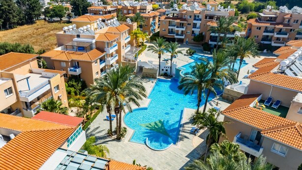 A residential area with a swimming pool surrounded by palm trees and apartment buildings with orange-tiled roofs. - Iris Village 201 (Paphos)