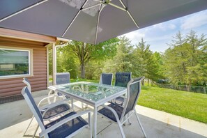 Eine Terrasse im Freien mit Glastisch und -stühlen, einem großen Sonnenschirm und Blick auf eine Rasenfläche mit Bäumen.