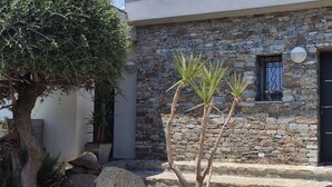 A stone building with a black door and windows, surrounded by a small garden with a tree and plants. - Studio à Mora Dell Onda 20137 Lecci (LECCI)