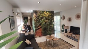 A modern living room with a large tropical mural, a black sofa, a TV, and a coffee table. - Studio à Mora Dell Onda 20137 Lecci (LECCI)