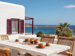 Une terrasse avec une table dressée pour le dîner, offrant une vue sur la côte, avec des bateaux et des collines en toile de fond.