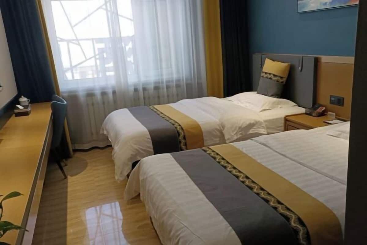 Une chambre d’hôtel avec deux lits, un bureau, une chaise, une table de chevet et une fenêtre avec des rideaux légers.