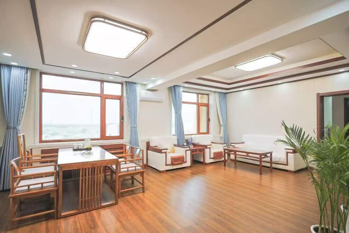 A spacious room with a wooden dining table and chairs, a sofa, and a small coffee table. There are large windows with curtains, and a potted plant in the corner.