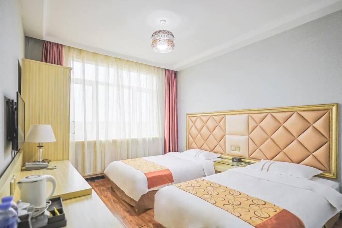 A hotel room with two beds, a bedside table with a lamp, a small table with a kettle, and a window with curtains.