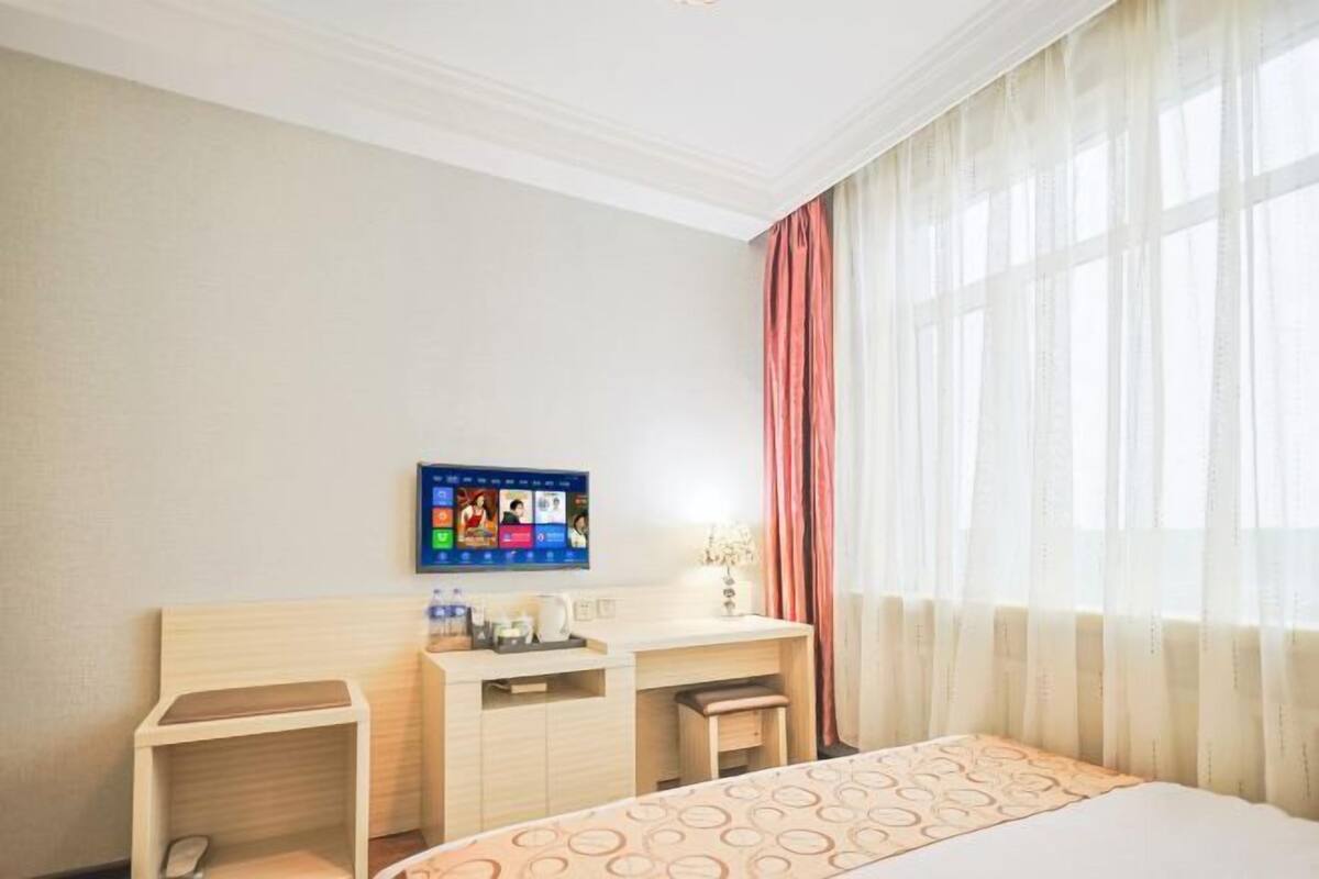 A hotel room with a bed, a desk, a chair, a TV mounted on the wall, and a window with sheer curtains.
