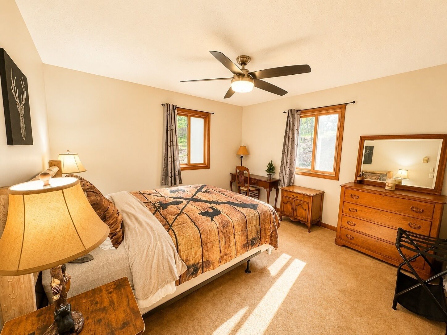 A bedroom with a bed, a desk, a dresser, a ceiling fan, and two windows with curtains.
