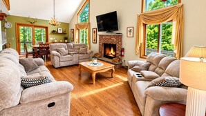 A spacious living room with a fireplace, comfortable seating, and large windows offering a view of greenery.
