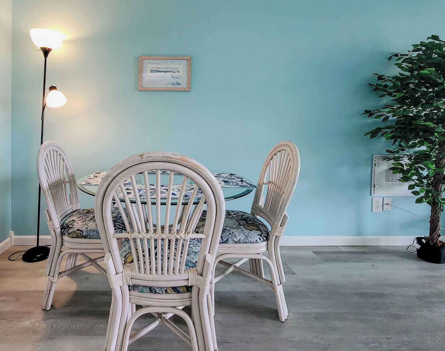 A dining area with a round table, two chairs, a floor lamp, a framed picture, and a potted plant.