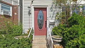 Une maison avec une porte rouge, une façade en briques et un petit jardin devant, agrémenté de plantes en pot.