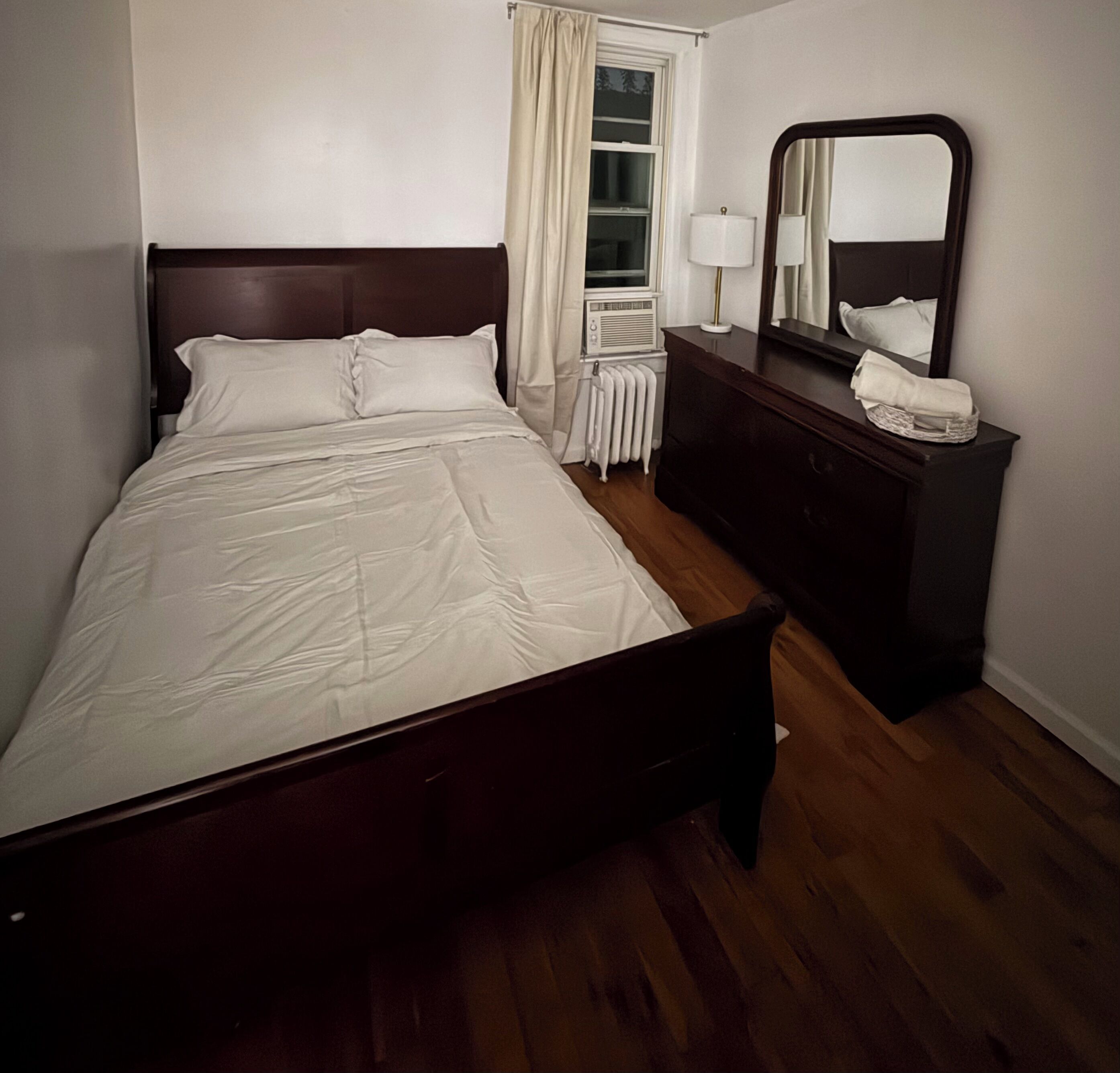 Una cama bien hecha con sábanas blancas, un cabecero de madera oscura, un aparador con espejo y un radiador en el suelo.