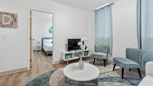 A modern living room with a round coffee table, a sofa, a TV stand, and a bed in the background. - Town St Medical Lofts (Columbus)