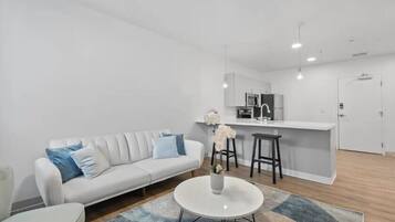 Een moderne woonkamer met een witte bank, een ronde salontafel, en een keukenhoek met een wit aanrecht en barstoelen.