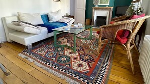 A living room with a white sofa, a glass coffee table, a patterned rug, a wicker chair, and a fireplace. - L'appartement Jean Moulin  (Paris)
