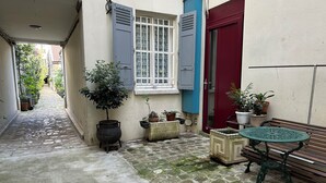 A narrow alleyway with a cobblestone path, a potted tree, a black cauldron, and a wooden bench with a green table. - L'appartement Jean Moulin  (Paris)