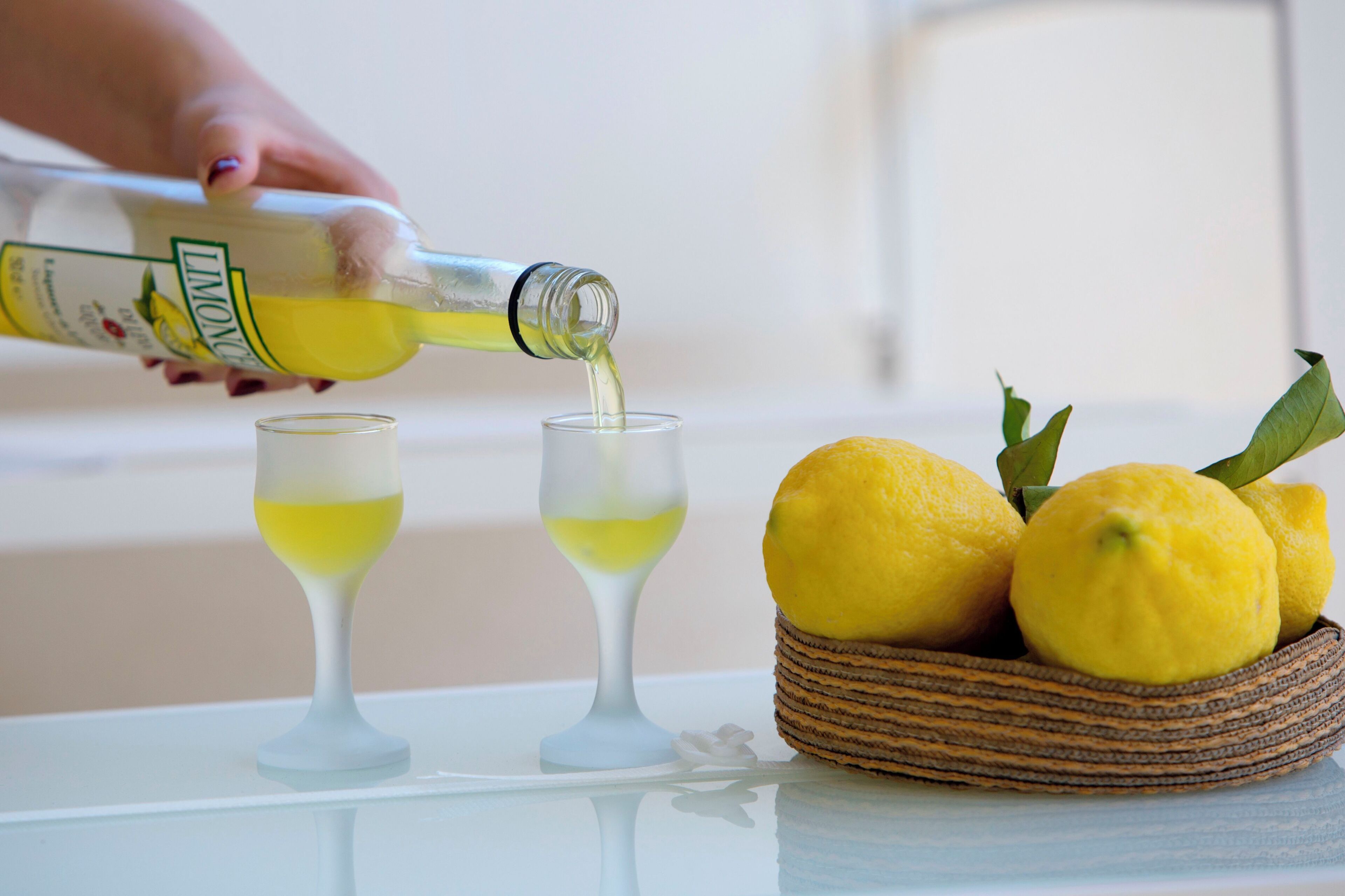 Une main verse un liquide jaune d’une bouteille dans deux verres. On voit des citrons dans un panier à côté des verres.