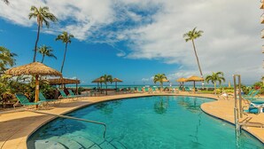 A pool area with lounge chairs and umbrellas, surrounded by palm trees, with a clear view of the ocean. - Top floor oceanfront penthouse w/ wrap-around lanai and pool (Lahaina)