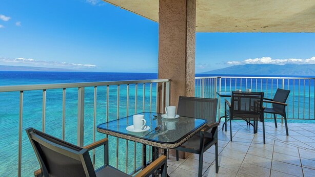 A balcony with a glass table and chairs, overlooking a clear blue sea and distant mountains. - Top floor oceanfront penthouse w/ wrap-around lanai and pool (Lahaina)