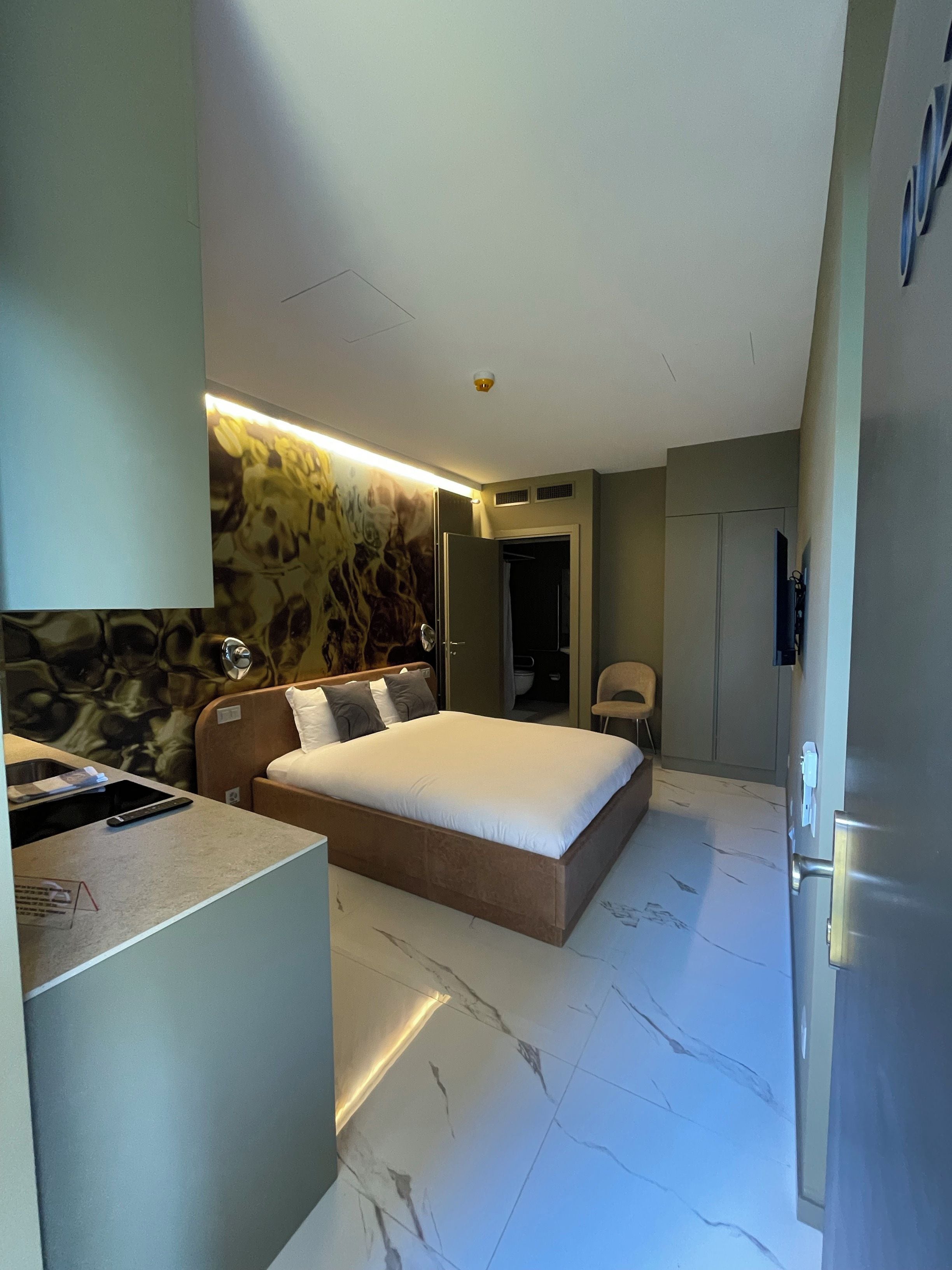 A compact hotel room with a bed, bedside table, and a small desk area.